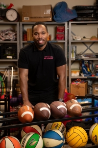 Man standing in front of footballs and basketballs with boxes behind him.